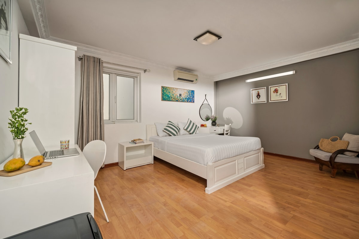 A studio apartment features a spacious bed with accent pillows against a neutral backdrop. Natural light enters through a window, illuminating a simple desk with a chair and decorative items. A cozy seating area is present, complemented by light wood flooring and soft color tones.