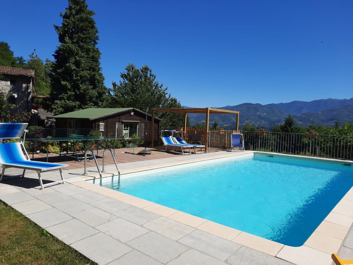 An outdoor area features a private swimming pool surrounded by paved patio. Lounge chairs with vibrant blue fabric are arranged nearby. In the background, a wooden structure provides shaded seating, with manicured greenery and mountain views completing the scene.