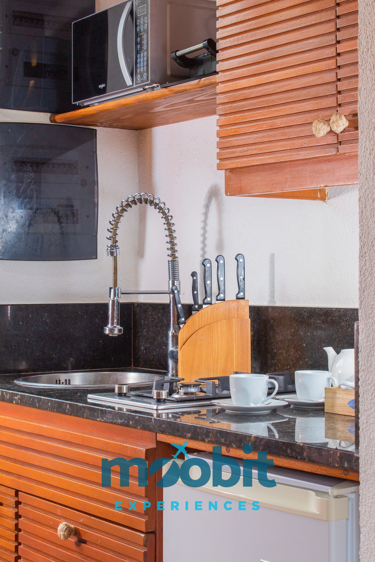 A compact kitchen area features wooden cabinetry and a dark granite countertop. Kitchen utensils are organized on the counter, while a set of dishes and a kettle are neatly arranged. A microwave is positioned above the countertop, contributing to the functional design.