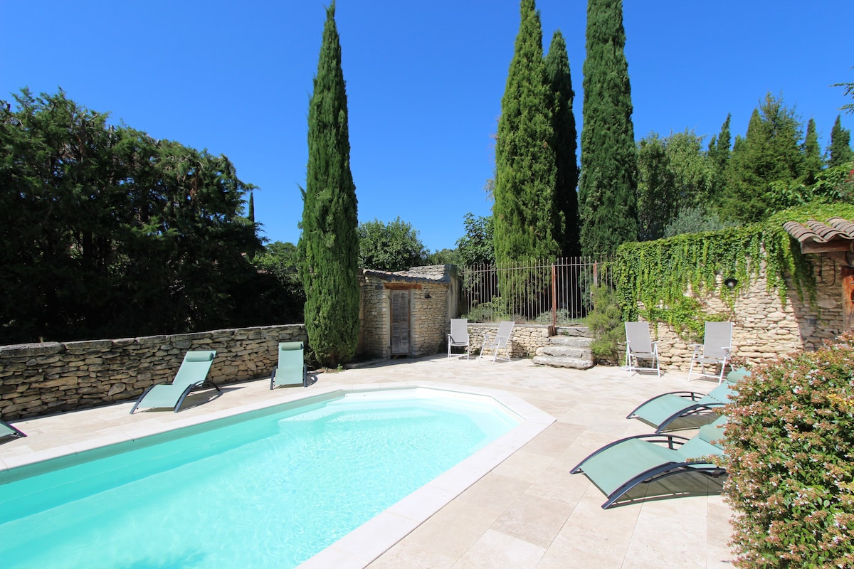 A clear water swimming pool is set within a stone courtyard, surrounded by neatly trimmed cypress trees. Several lounge chairs are positioned around the pool area, along with a stone seating arrangement, creating inviting spaces for relaxation.