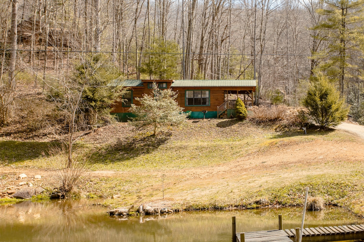 A charming cabin is set amidst a natural landscape, with a green metal roof blending into the surrounding trees. The cabin overlooks a small pond, while a gentle slope leads down to the water's edge, enhancing the serene rural environment.