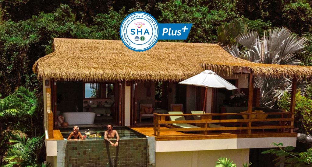 A tropical cottage is nestled among lush greenery, featuring a thatched roof and open design. A private pool with natural stone tiles extends from the terrace, which is furnished with comfortable seating and an umbrella for shade. Two guests are seen enjoying the serene environment.