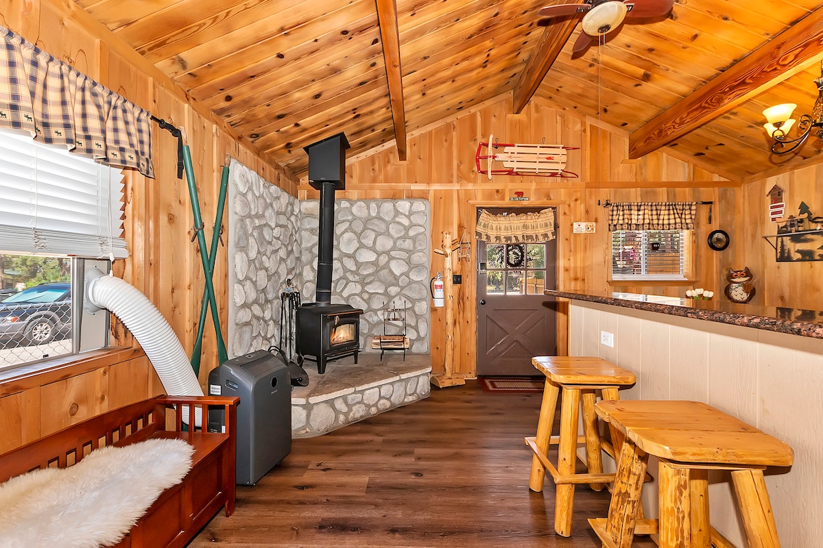 A rustic cabin interior showcases a stone fireplace as the focal point, complemented by wooden beams and paneling. Two wooden stools are positioned near a bar area, with a plush seating option and a portable air conditioning unit visible. Natural light enters through a window adorned with matching valance.