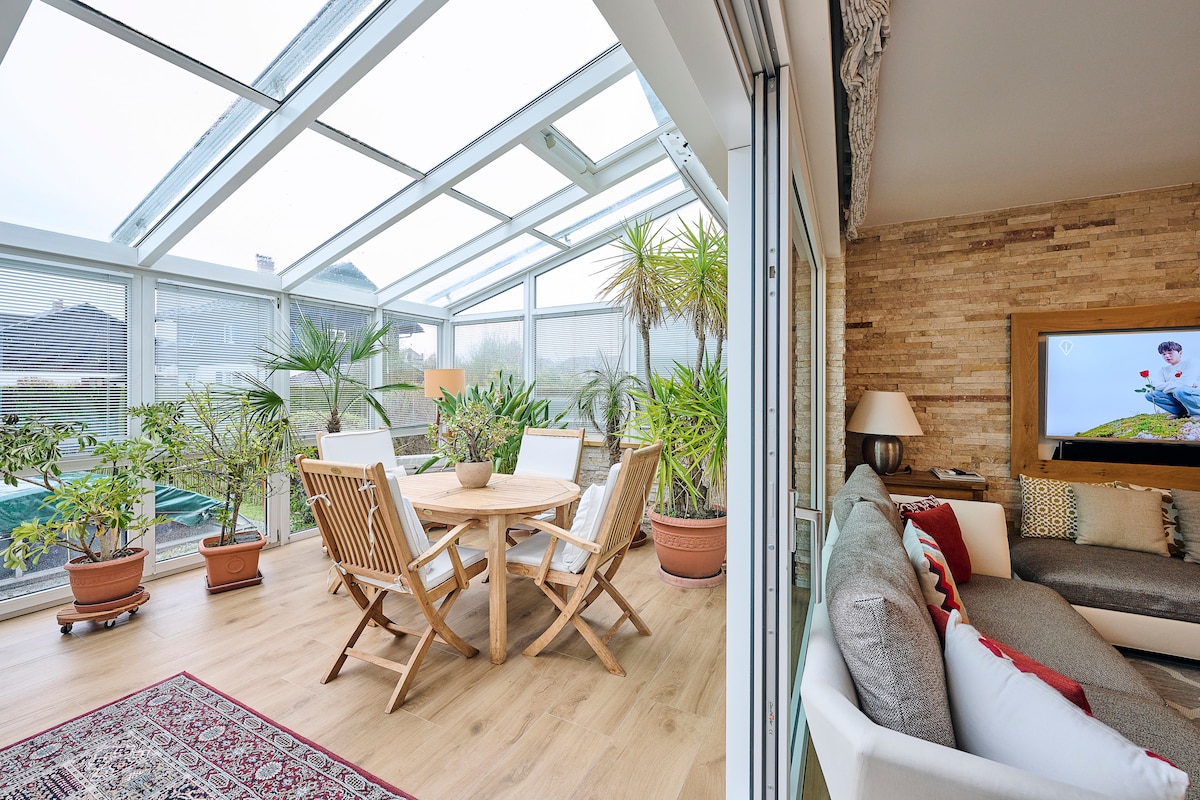A bright garden room features large glass panels allowing natural light to fill the space. A wooden round table is surrounded by five deck chairs, while potted plants add greenery to the ambiance. A cozy seating area is visible, enhancing the overall inviting atmosphere.