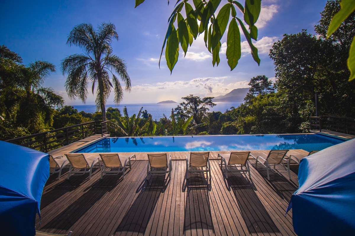 An outdoor pool area is surrounded by lush greenery, offering views of distant mountains and the sea. Eight lounge chairs are positioned neatly along the poolside, with shades providing comfort. The scene is set under a bright sky, indicating a pleasant and sunny day.