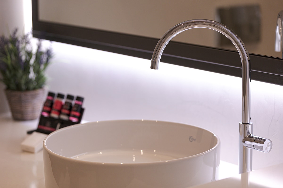 A modern bathroom sink is featured, presenting a round white basin complemented by a sleek chrome faucet. A backlit mirror enhances the space's brightness, while a small potted plant adds a touch of greenery. A selection of toiletries is neatly arranged beside the sink.