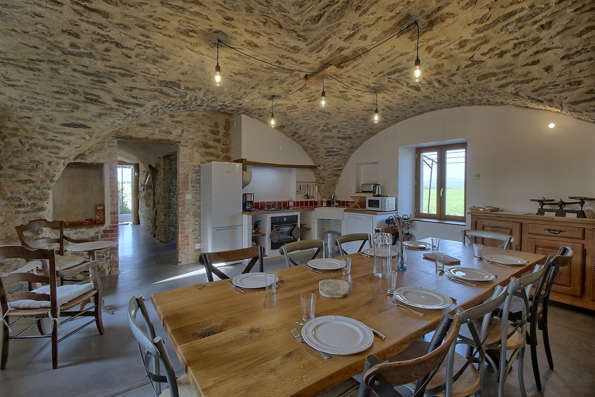 An open dining area features a large wooden table surrounded by metal and wooden chairs. Industrial-style lighting illuminates the rustic stone walls and ceiling. A modern kitchen area is visible, equipped with essential appliances, and a window offers views of the outdoor landscape.