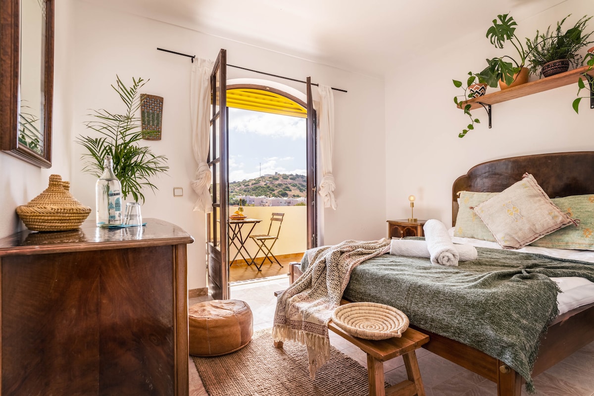 A cozy bedroom features a spacious bed adorned with decorative pillows and a soft blanket. Double doors open to a private balcony, revealing a small outdoor table and chairs. Natural light fills the room, complemented by greenery and wooden accents.