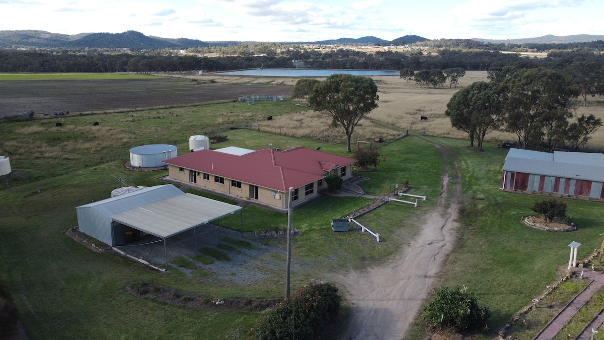 An aerial view showcases a spacious home with a red roof, surrounded by green lawns and open fields. The property features outbuildings, water tanks, and a circular drive. In the background, rolling hills and a tranquil body of water are visible, encapsulating the serene rural setting.