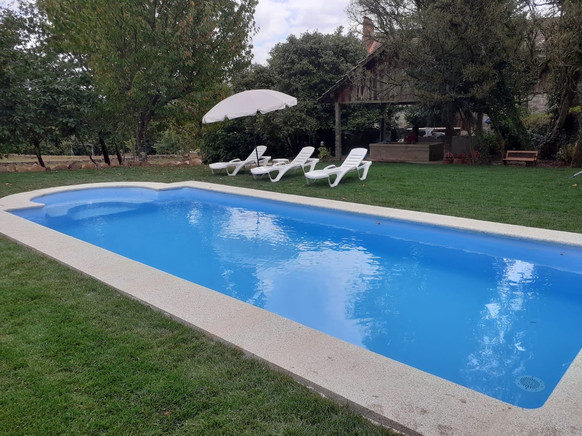 A private swimming pool with clear blue water is surrounded by green grass and sun loungers. An umbrella provides shade nearby, and trees are visible in the background, enhancing the outdoor space for relaxation and leisure.