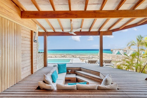 An inviting seating area features a sunken lounge surrounded by soft cushions in shades of white and turquoise. The space opens to a view of the ocean, with a private pool visible in the background, framed by a wooden deck and palm foliage.