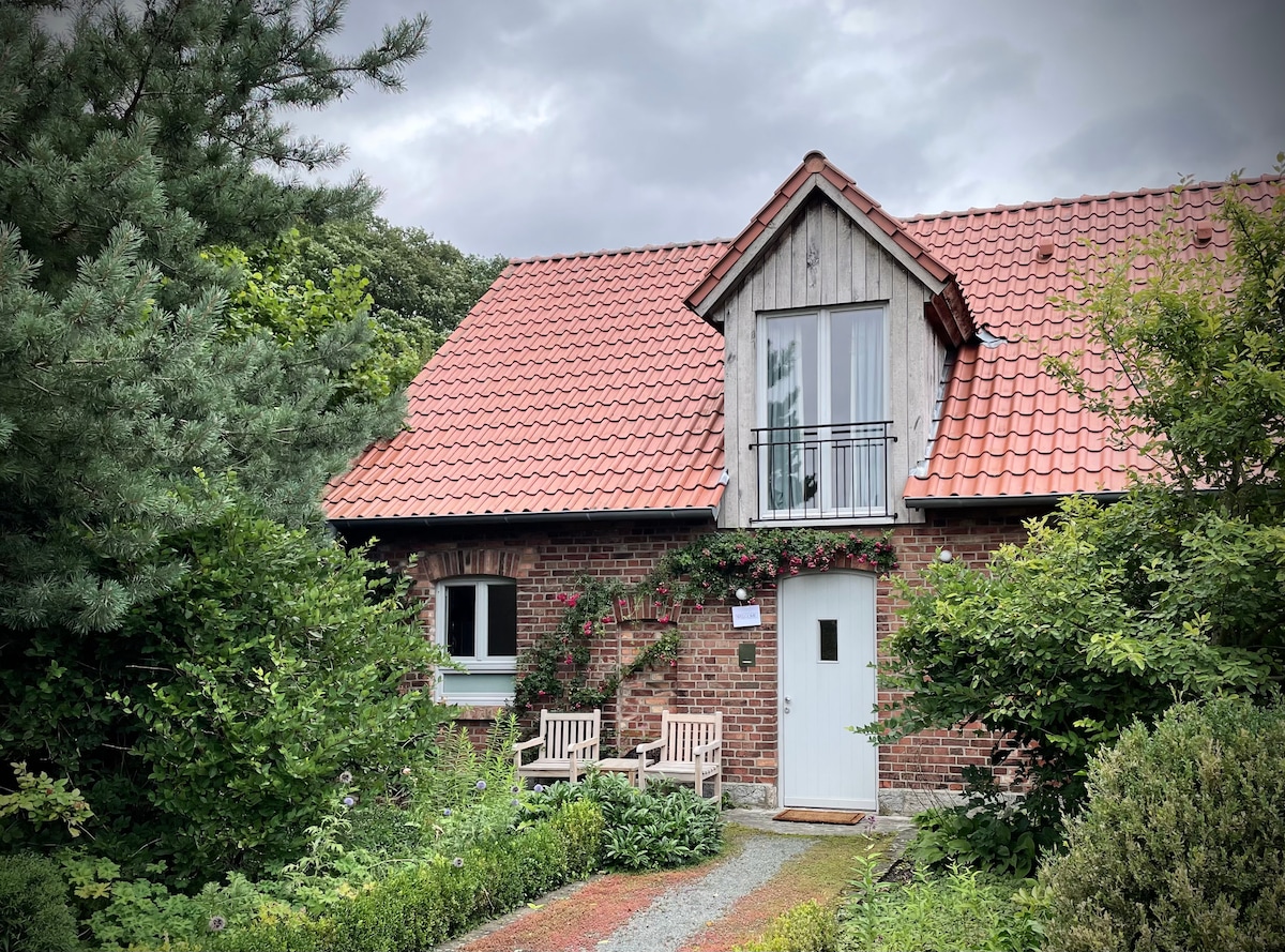 The exterior of a charming brick house is framed by lush greenery and decorative climbing plants. A small pathway leads to a white door under a sloped roof, accented by a balcony above. Two wooden benches are positioned on either side of the entrance.