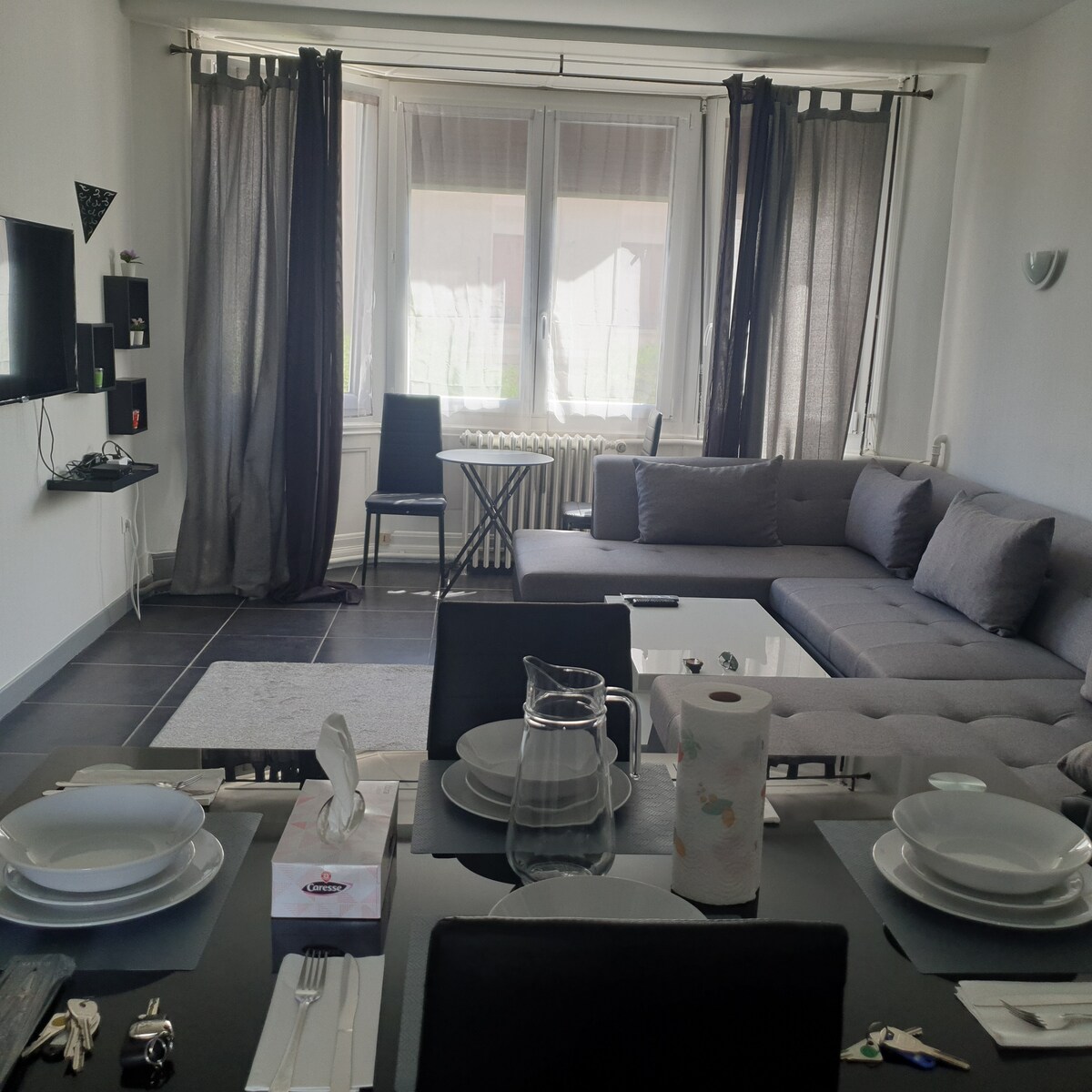A living area is presented with a large gray sectional sofa and a glass coffee table. A dining table is set with plates and cutlery. Large windows are covered with light curtains, allowing natural light to brighten the space. A wall-mounted TV is visible.