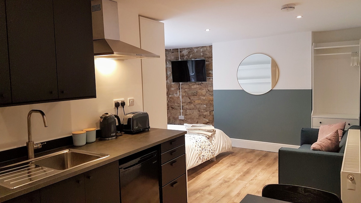 A modern studio flat features a kitchenette with sleek cabinetry and stainless steel appliances. The lounge area includes a comfortable sofa and a wall-mounted television. A spacious bed is located in the background against a partially exposed stone wall, complemented by circular mirrors.