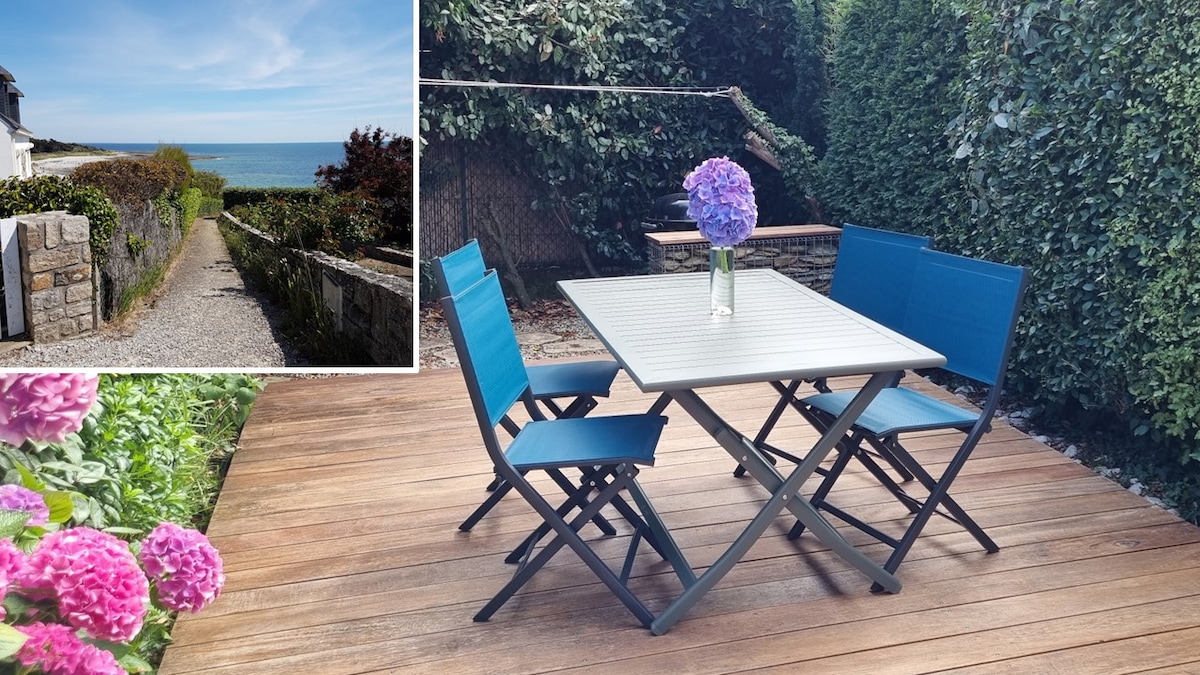 A wooden terrace is equipped with a dining table and four folding chairs, surrounded by lush greenery. A path leading to the coastline is visible in the inset, providing a glimpse of the nearby sea and blooming flowers in the foreground.
