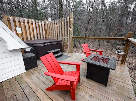 An outdoor deck is featured, surrounded by a wooded landscape. Two red adirondack chairs are positioned near a fire pit, which is set on a wooden surface. A hot tub is located at the back, enclosed by wooden fencing and soft ambient lighting.