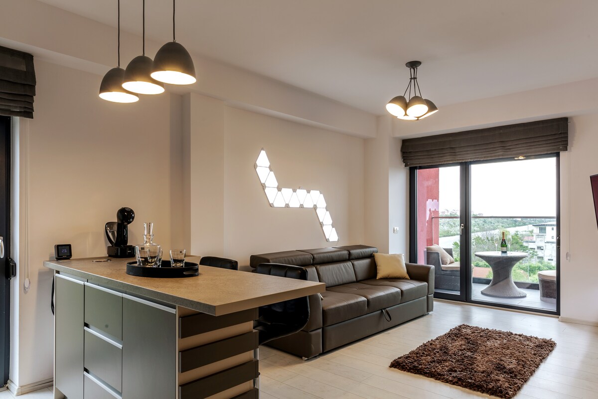 The living area features a spacious sofa facing large glass doors that open to a balcony, offering views of the surrounding landscape. A modern kitchen island with bar seating is positioned nearby, while decorative wall lights provide subtle illumination.