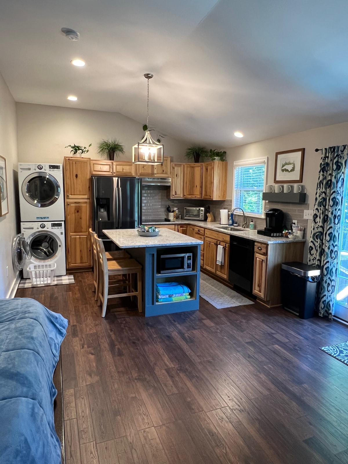 The fully furnished kitchen features wooden cabinetry and a large island with seating for two. Appliances include a refrigerator, microwave, and coffee maker. A washer and dryer are located nearby. Natural light filters in through the window, complemented by tasteful decor.