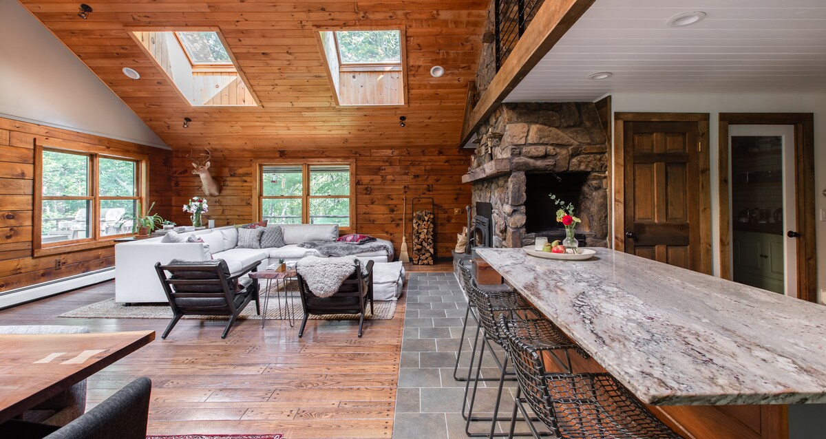 A modern log cabin living area features warm wooden walls and large skylights, allowing natural light to fill the space. A cozy seating arrangement includes a sectional sofa with textured blankets. A stone fireplace adds a rustic touch, while a sleek kitchen island showcases stylish bar stools.