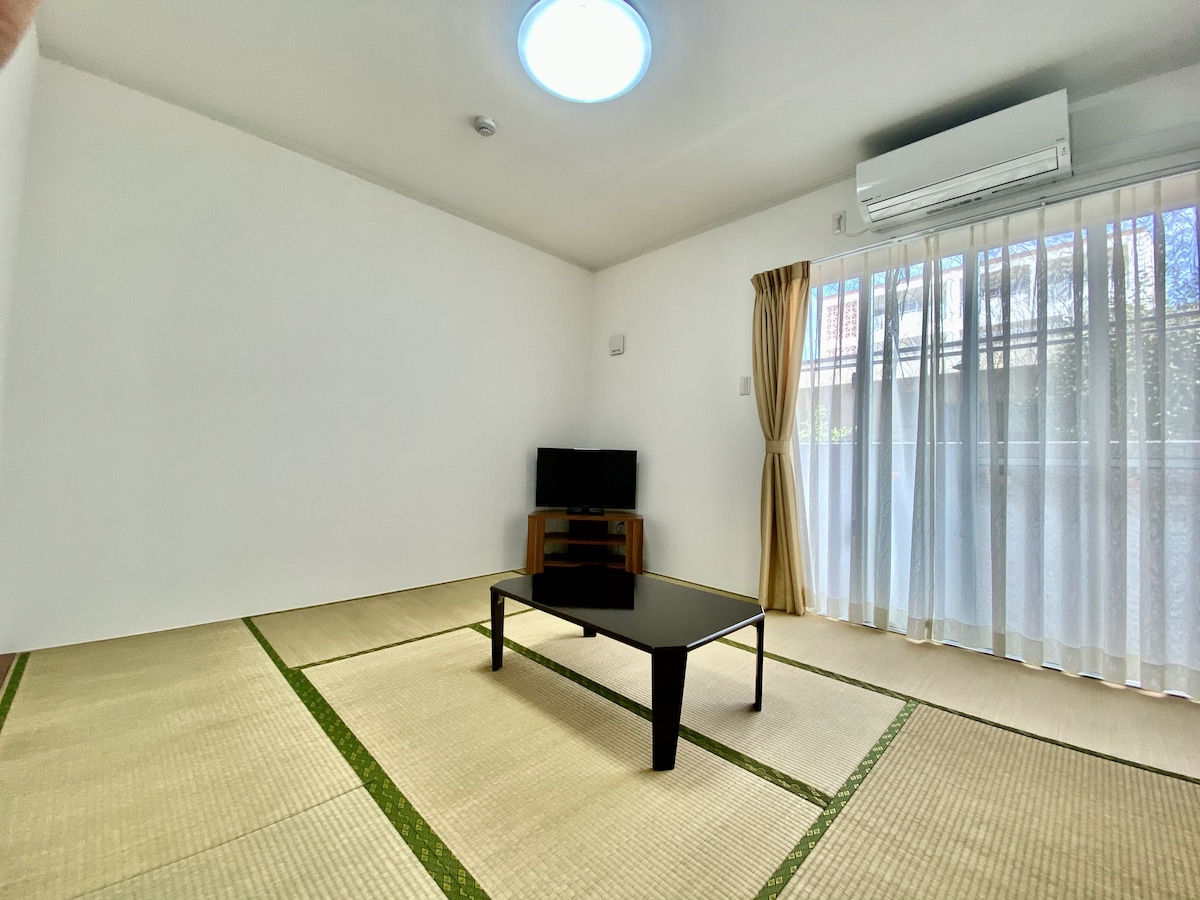 A spacious room is displayed with tatami flooring, featuring a low black coffee table at the center. Sunlight filters through sheer curtains, illuminating the area. A television is positioned against the wall, while a round ceiling light provides additional brightness.