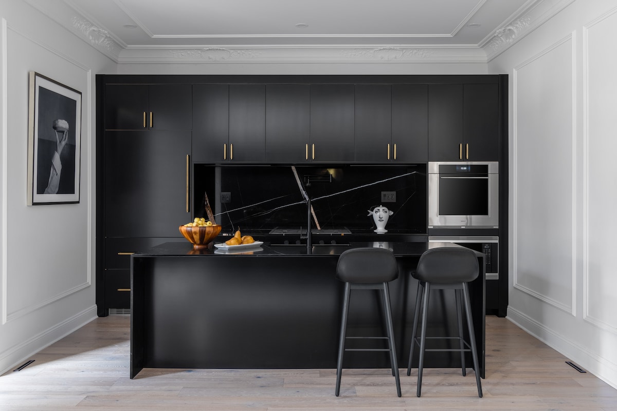 A modern kitchen is displayed, featuring sleek black cabinetry with gold hardware. A central island with bar stools provides seating. In the background, high-end appliances, including a wall oven, are visible. A bowl of fruit adds a touch of color to the countertop.
