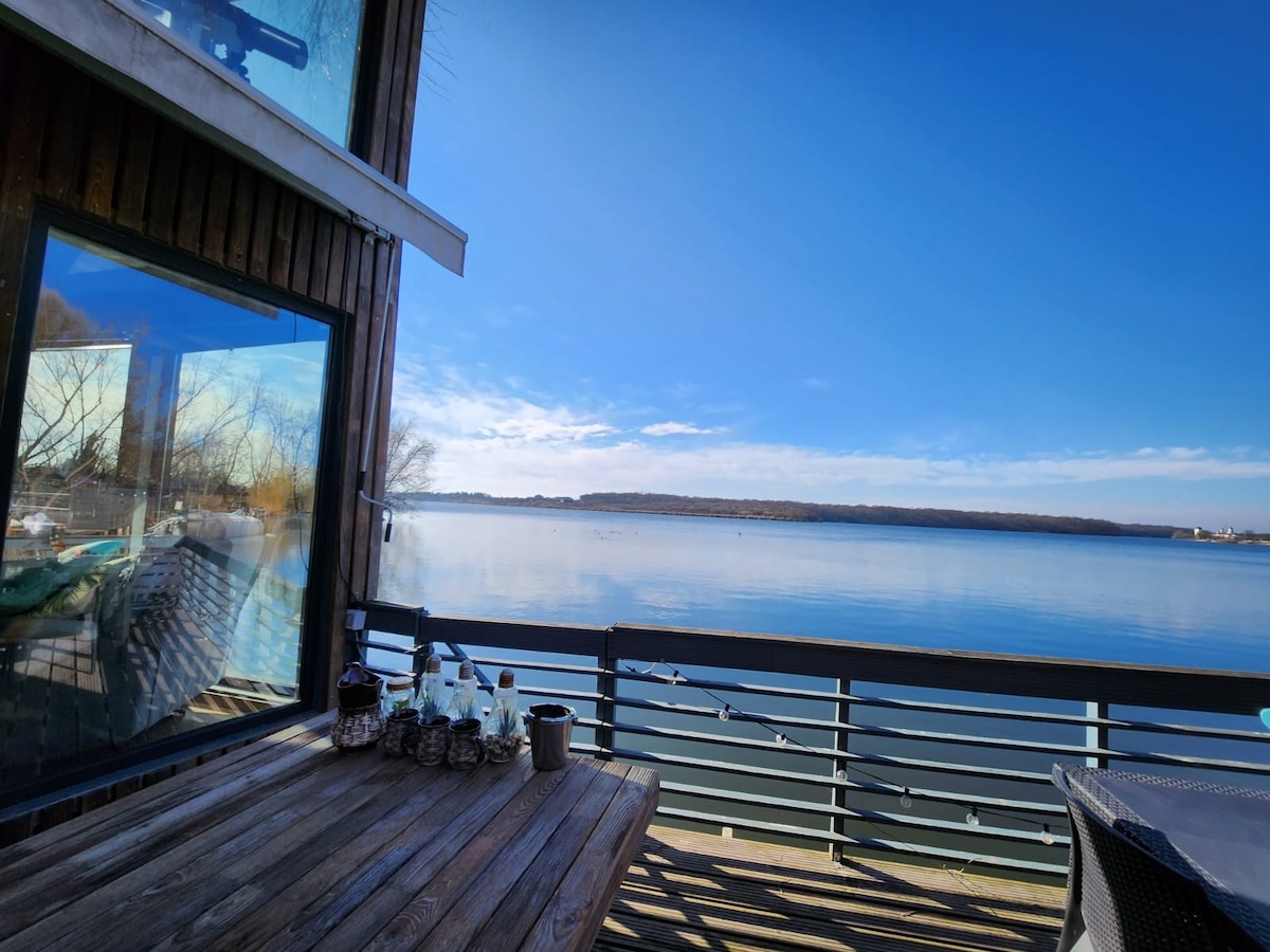An outdoor dining area features a wooden table overlooking a serene lake. Large glass windows provide expansive views of the water, reflecting the clear blue sky. Subtle decorative elements are arranged on the table, enhancing the relaxing ambiance of the space.