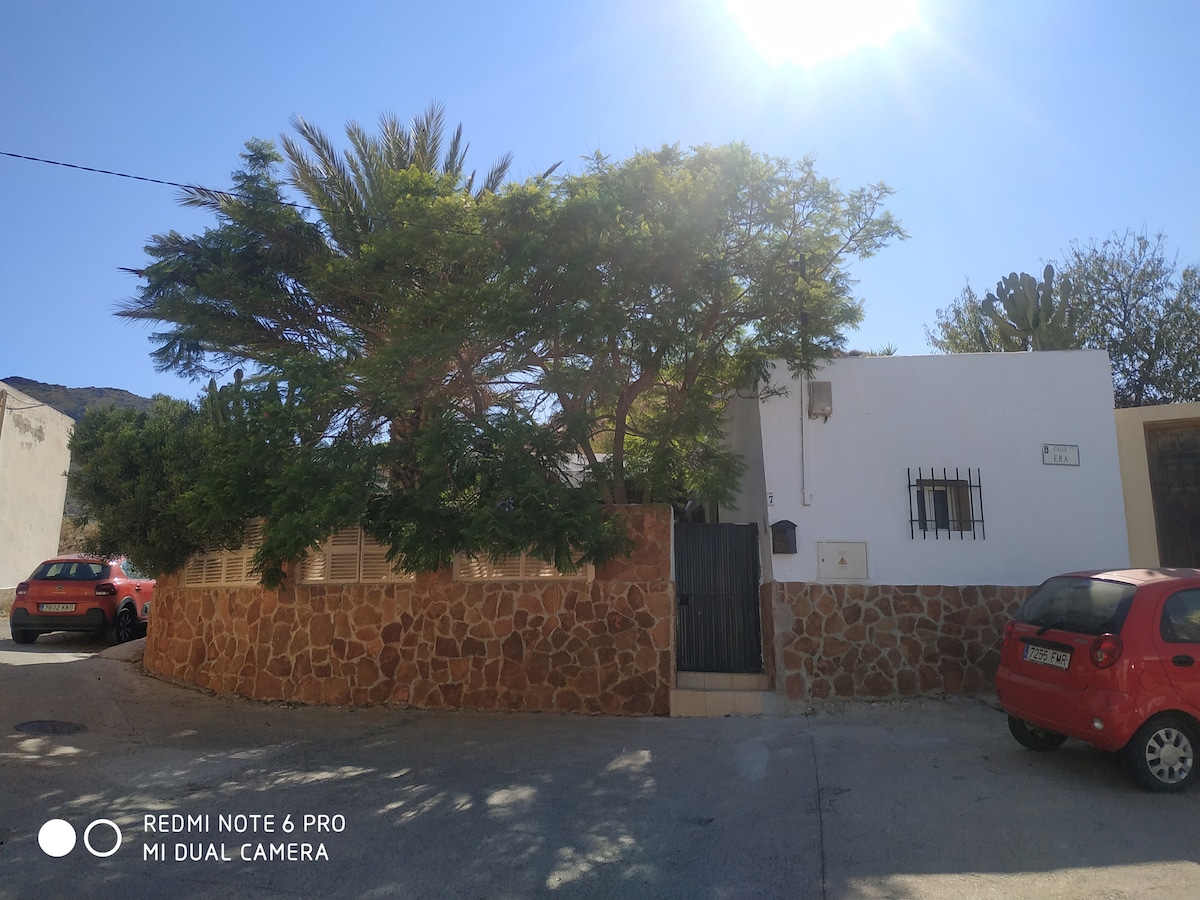 The exterior view features a white building surrounded by a stone wall, with lush greenery providing shade. A single entrance gate is visible, framed by a textured stone façade. The area is bathed in sunlight, highlighting the surrounding landscape.