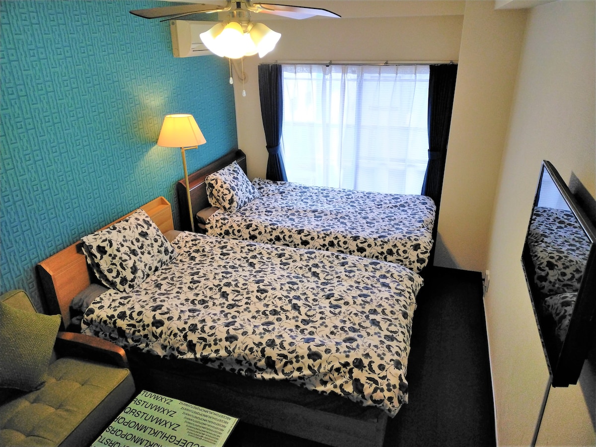 The sleeping area contains two spacious single beds adorned with floral bedding. A lamp stands next to a window, which allows natural light to illuminate the room. A small sofa is positioned in one corner, enhancing the inviting ambiance.