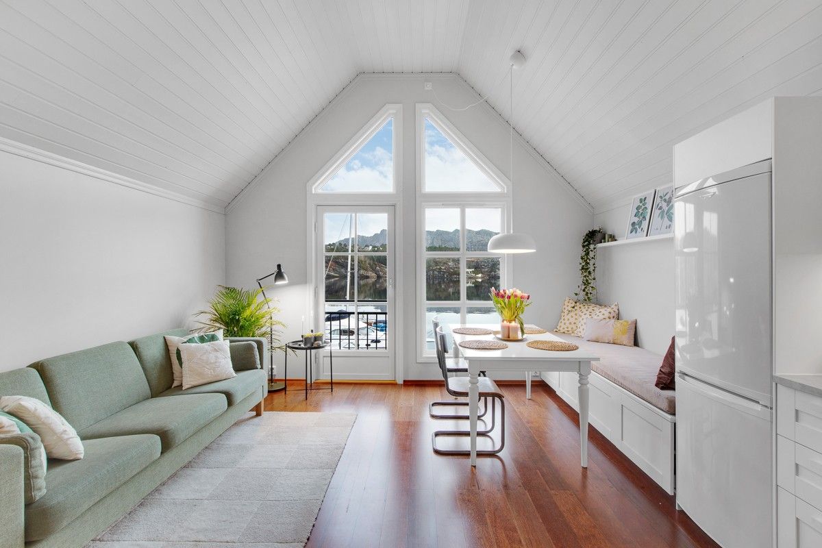 A bright living space features large triangular windows offering views of the fjord. A light green sofa is positioned adjacent to a white dining table surrounded by metallic chairs. Natural light fills the room, accentuating the wooden floor and minimalistic decor.