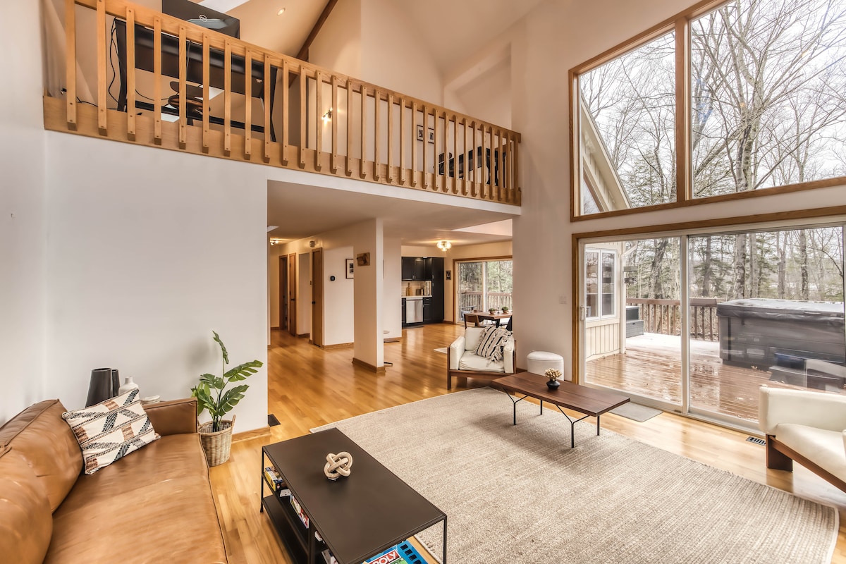 An open living area features a leather sofa facing a stylish coffee table, situated on a light-colored area rug. Large windows allow natural light to fill the space, highlighting the wood accents and vaulted ceiling. Access to the deck and hot tub is granted through sliding glass doors.