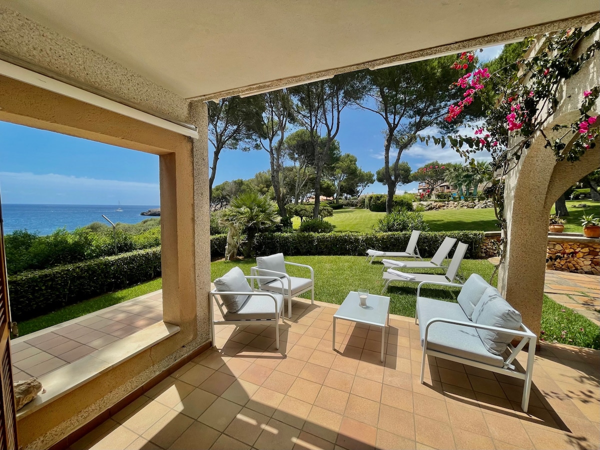 A private terrace is visible, featuring white lounge chairs and a small table. The sea can be seen in the background, framed by lush greenery and trees. The area offers a peaceful outdoor setting, ideal for relaxation and enjoyment of the surrounding nature.