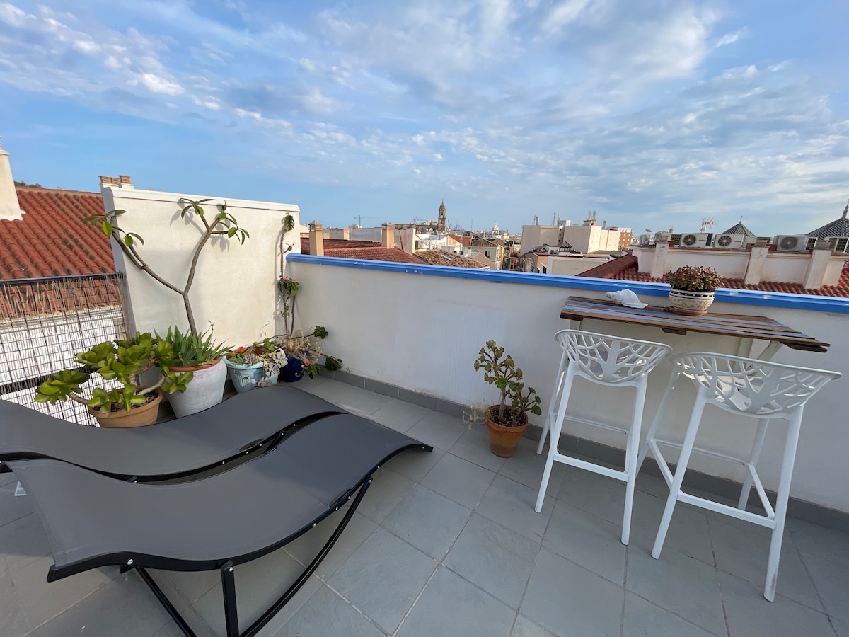A rooftop terrace is presented with a lounge chair and a small dining table. Potted plants add greenery to the space, while a view of the cityscape and distant church tower can be seen against the sky. Light grey tiles cover the terrace flooring.