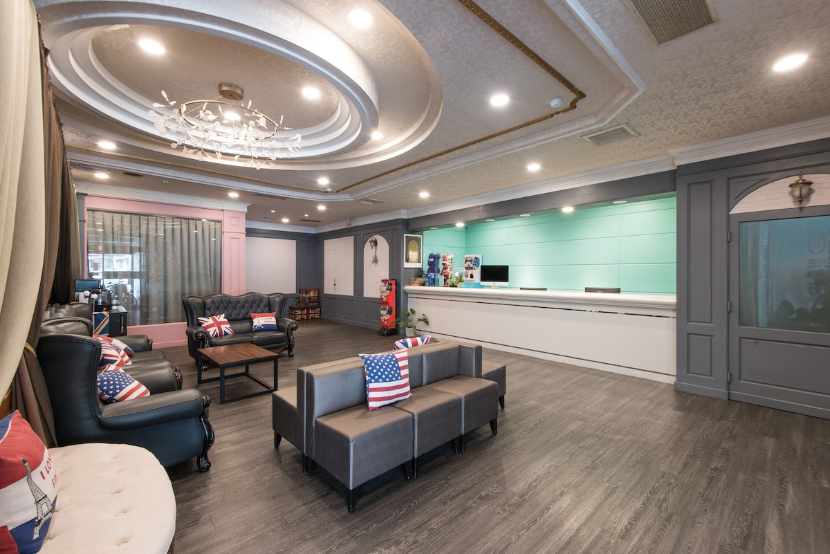 The lobby area features a spacious reception desk with a light blue backdrop. Comfortable seating arrangements include a black leather sofa and a gray fabric couch, adorned with decorative pillows. A chandelier hangs from the ceiling, while soft lighting enhances the welcoming environment.
