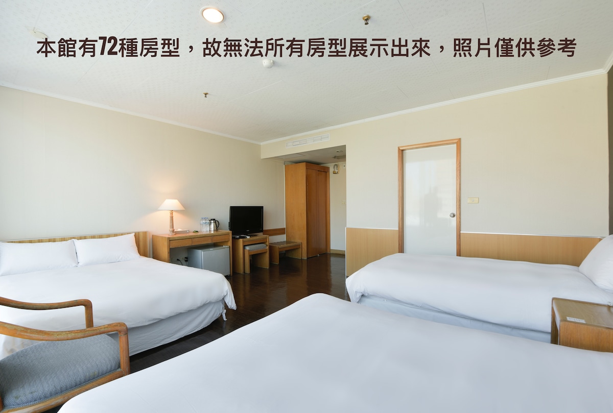 A spacious guest room is presented with two large beds positioned against light-colored walls. A wooden table and chairs are situated in the corner, and large windows allow natural light to fill the area. Neutral tones create a calm and welcoming environment.