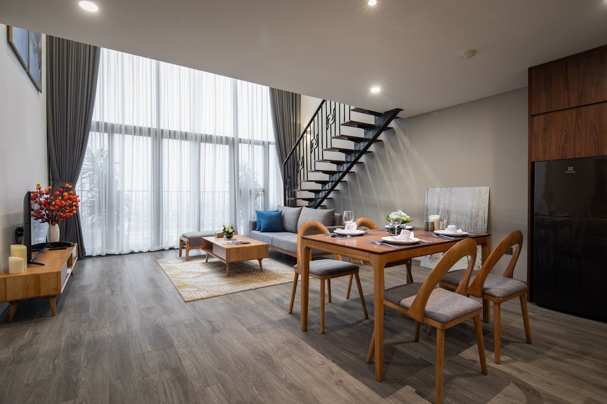 A spacious living area is highlighted by large windows allowing natural light to flow in. A dining table with wooden chairs accommodates several guests, while a staircase features sleek metal railings. A cozy seating area invites relaxation, complemented by tasteful decor and a warm wooden floor.
