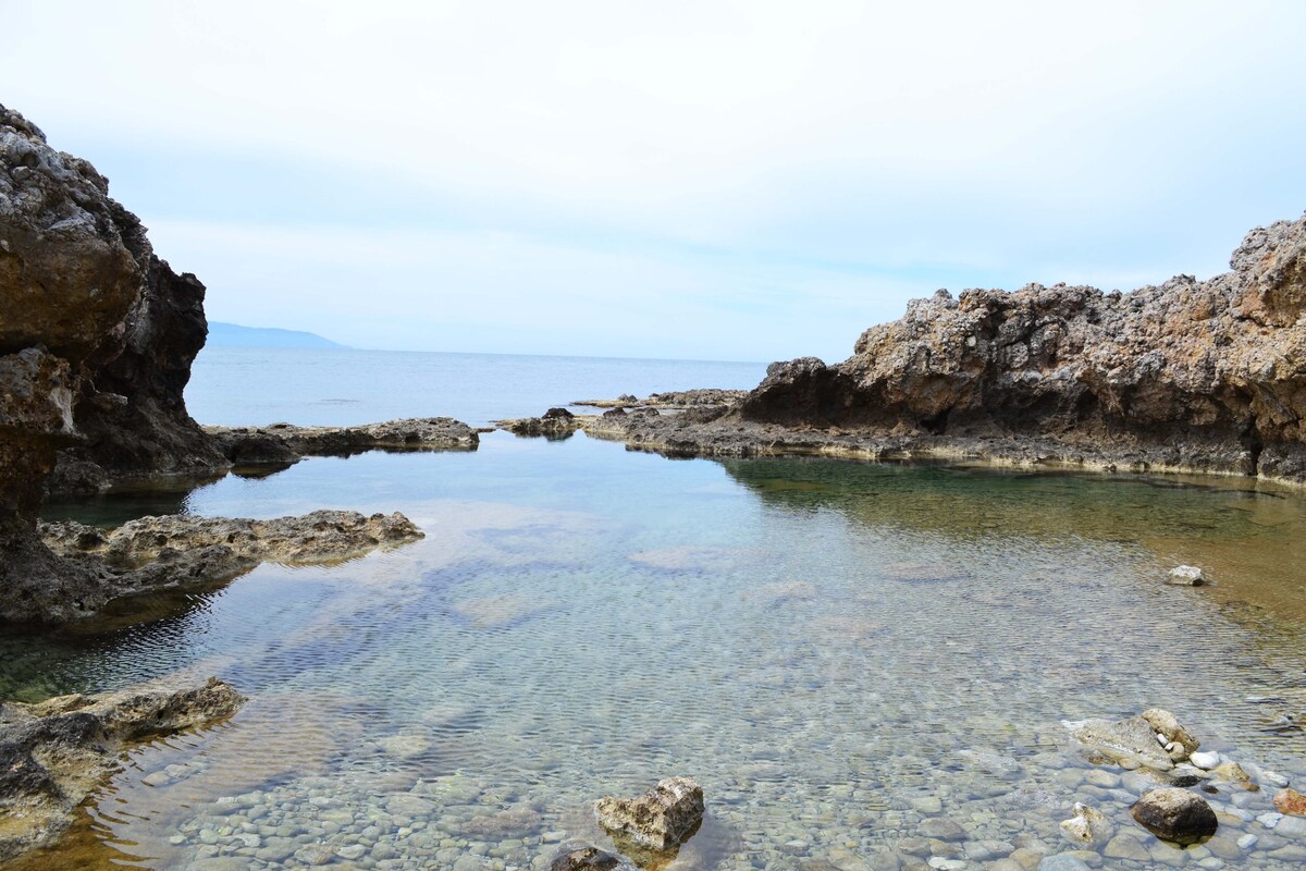 A tranquil rocky pool is surrounded by rugged cliffs, with clear water revealing pebbles and small stones beneath the surface. The horizon presents a serene view of the sea and distant land, creating a peaceful and natural setting.