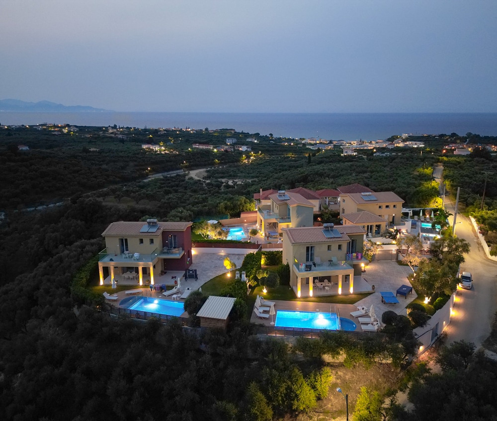 An aerial view captures the cluster of modern villas illuminated at dusk, showcasing private pools surrounded by lush greenery. The expanse of the Ionian Sea is visible in the background, with twinkling lights from nearby areas adding to the serene atmosphere.