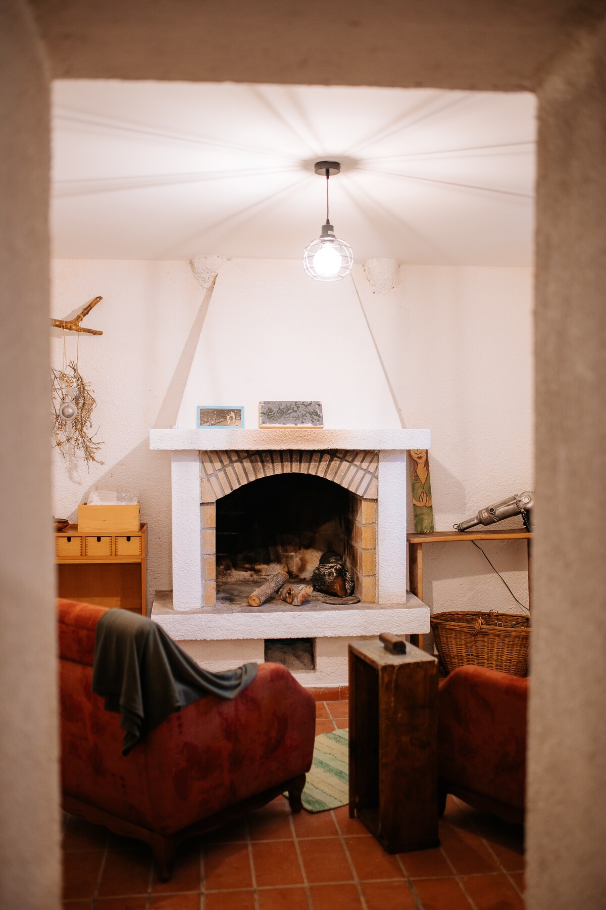 A cozy living area is framed by an archway, featuring a stone fireplace at its center. Two comfortable armchairs are positioned nearby, with a soft blanket draped over one. A wooden side table and a rustic basket add to the inviting ambiance.