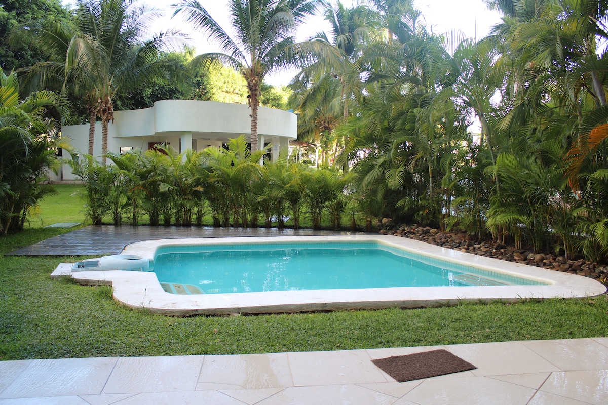 A private swimming pool is situated within a lush garden, surrounded by tropical palm trees. The clear blue water reflects the serene environment. Nearby, a deck area with wooden flooring complements the greenery, creating a tranquil space for relaxation.