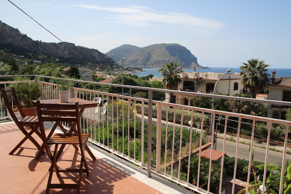 A spacious balcony offers a stunning view of the coastline, mountains, and sea. Outdoor seating is provided with wooden chairs and a small table, creating a serene space to enjoy the surrounding natural beauty.