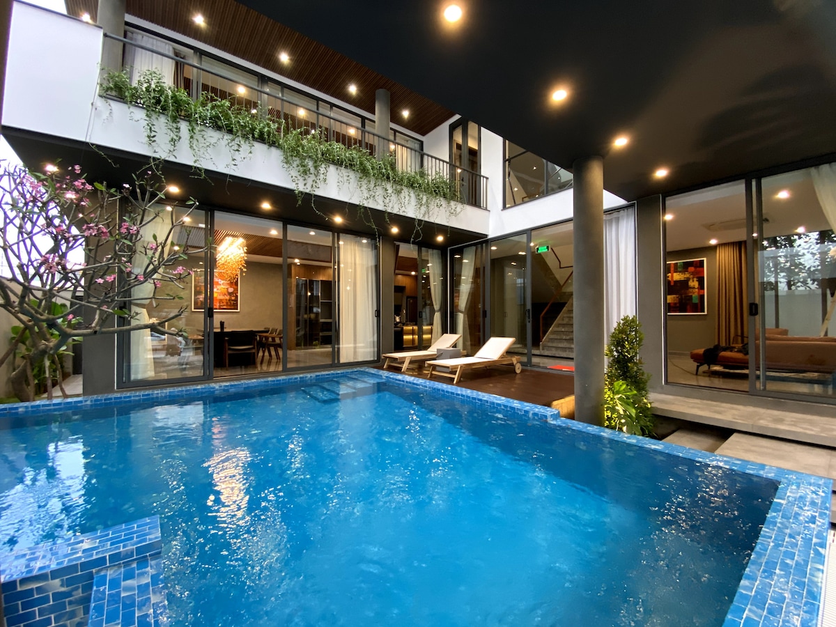 An inviting swimming pool is surrounded by a stylish outdoor area featuring lounge chairs. Floor-to-ceiling windows provide views of the living and dining spaces inside, complemented by lush greenery and soft lighting that enhance the tranquil ambiance.