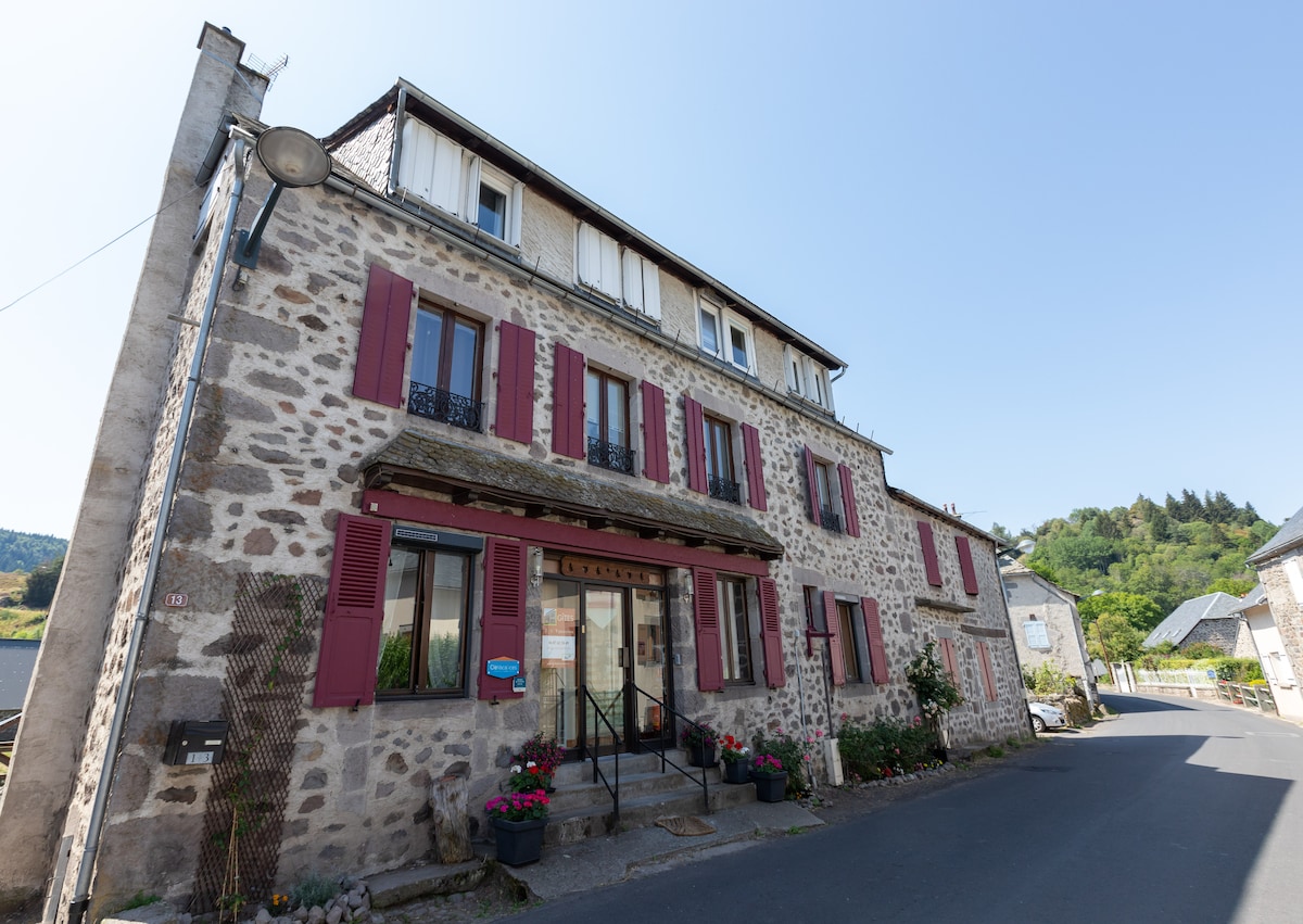 A charming stone building with red shutters and a tiled roof is located on a quiet street. Flower planters line the entrance, and the surrounding landscape features gentle hills, creating a peaceful setting. Sunlight highlights the building's rustic texture.