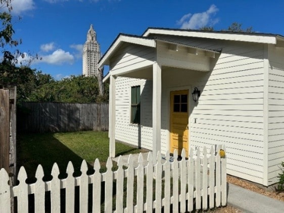 A quaint white cottage features distinctive architectural details, including a sloped roof and contrasting yellow door. A picket fence encloses the grassy yard, with trees providing a natural backdrop. The State Capitol building is visible in the distance under a clear blue sky.