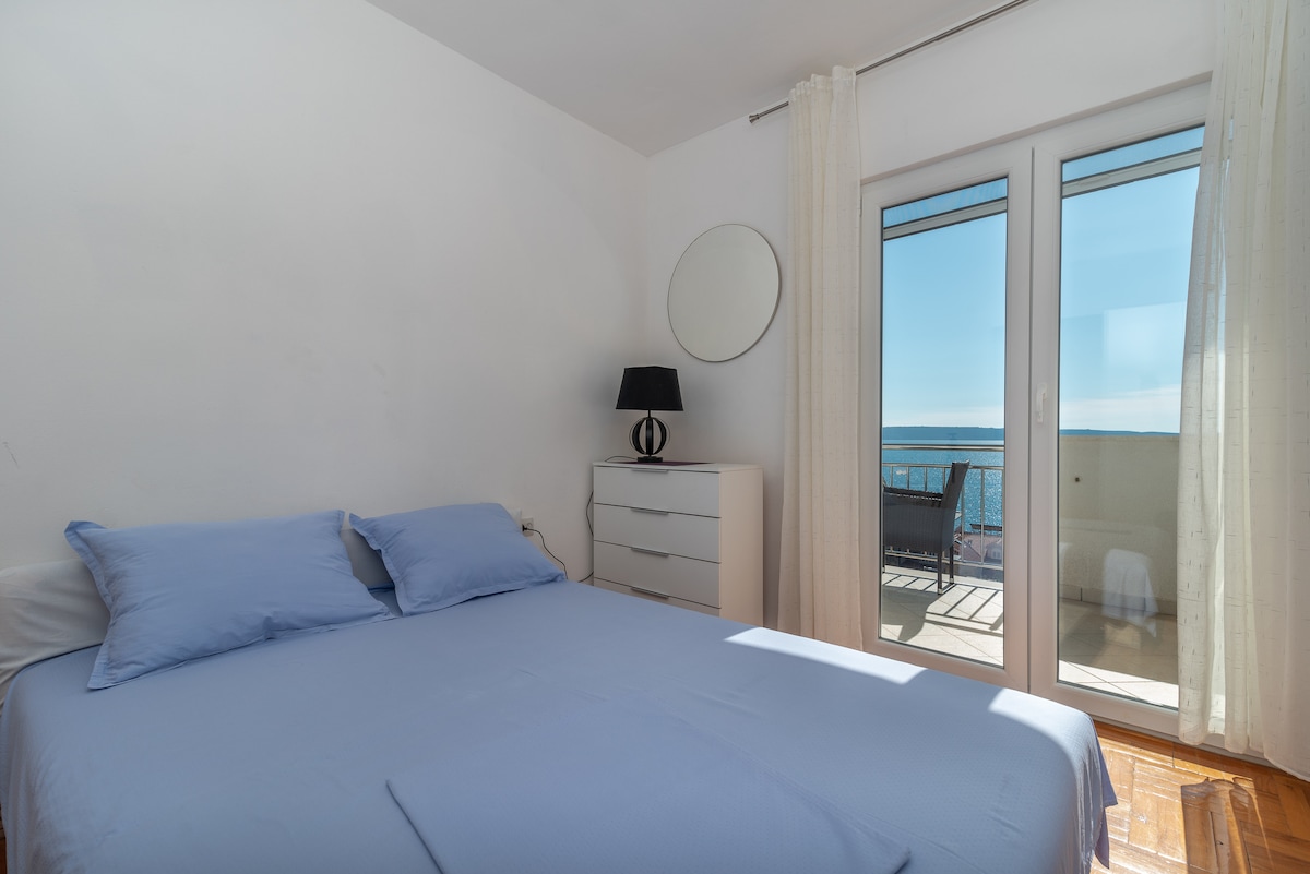 A bright bedroom features a spacious bed with light blue linens. A dresser stands against the wall, while a round mirror reflects natural light. Sliding glass doors open to a terrace with sea views, enhancing the serene ambiance of the room.