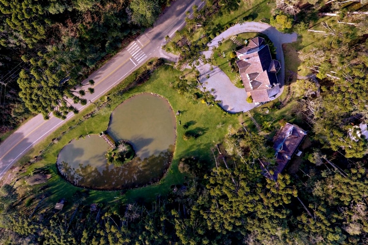 An aerial view captures the chalet and its surroundings, highlighting a heart-shaped lake and well-maintained gardens. The property is bordered by native forest, with a winding road visible nearby and ample green space for outdoor activities.