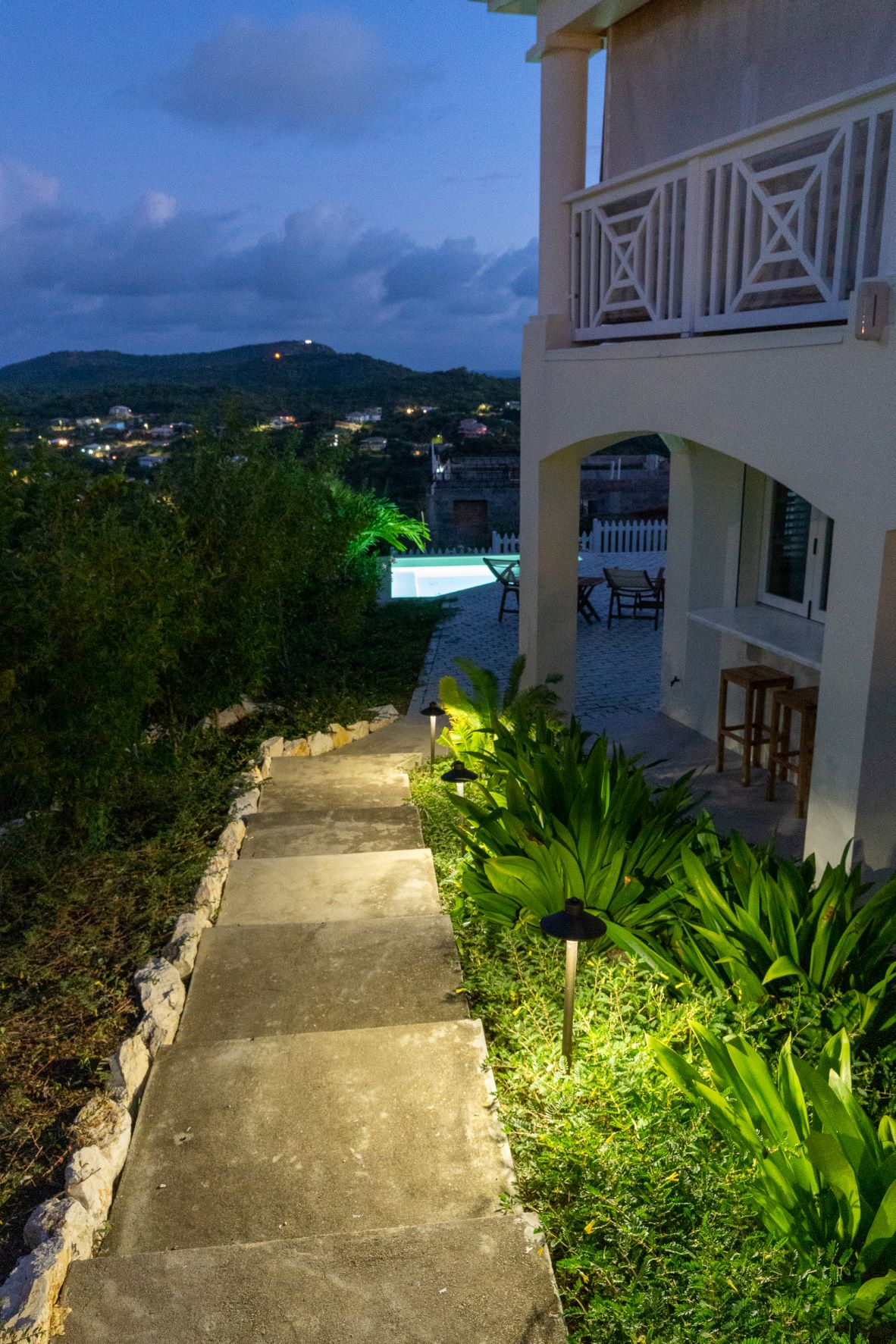A stone pathway is illuminated by soft landscape lights, leading from the lower garden area towards the main entrance. Lush green plants line the sides, while a distant view of the pool and hills can be seen under the evening sky.