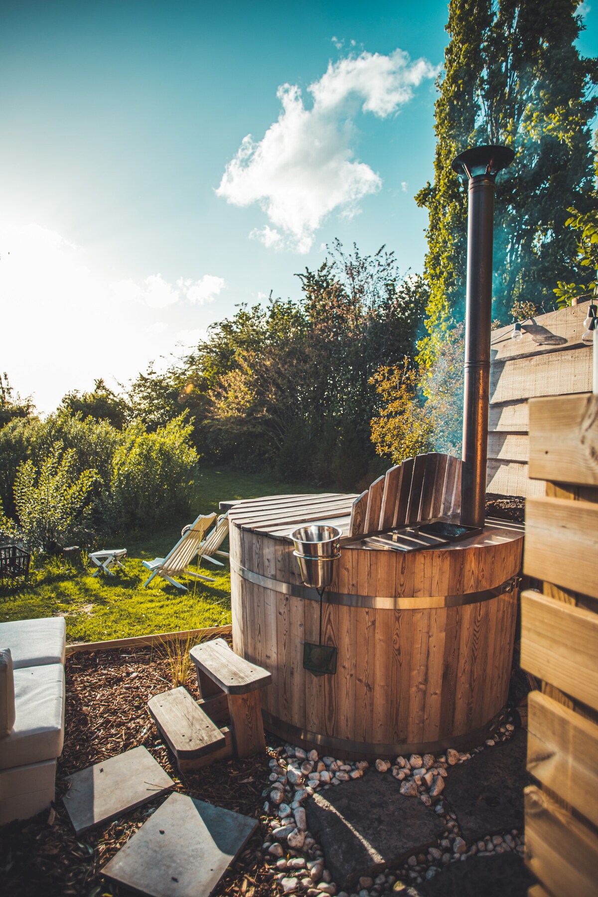 A wooden hot tub is positioned in a landscaped outdoor area, with a chimney releasing gentle smoke. Sunlight reflects off the tub, surrounded by greenery and stepping stones leading towards it. Lounge chairs are visible in the background, enhancing the serene natural setting.