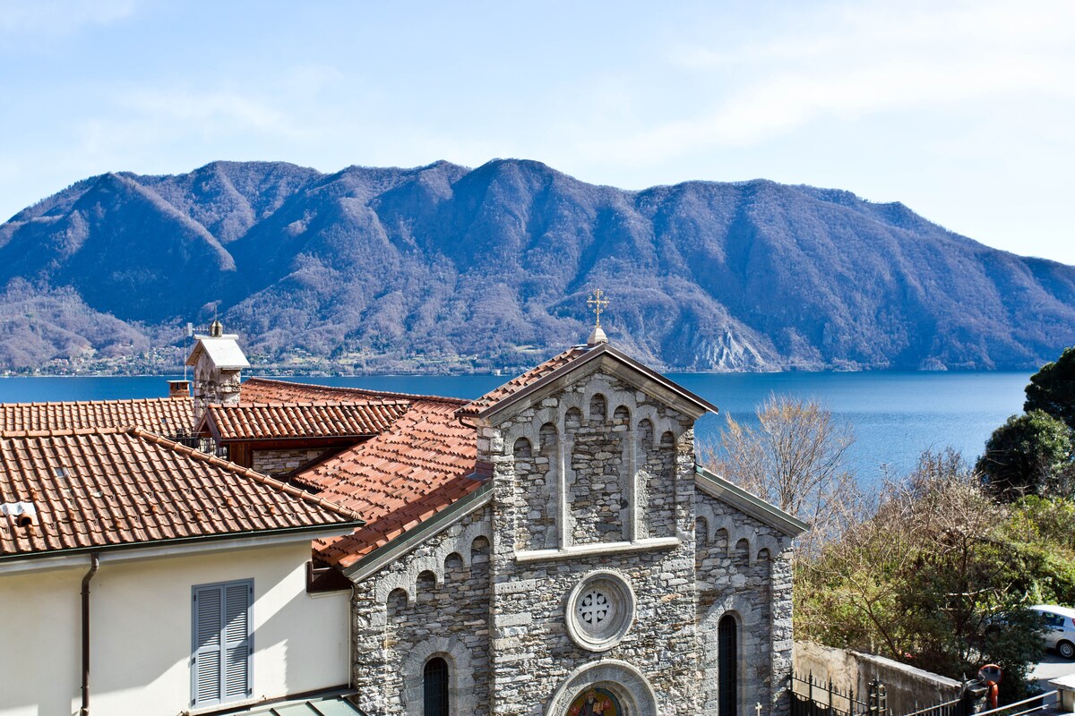 A scenic view encompasses a charming stone church with a terracotta roof, set against a backdrop of expansive mountains and a tranquil lake. The clear blue sky enhances the serene landscape, inviting peaceful reflection and appreciation of the natural surroundings.