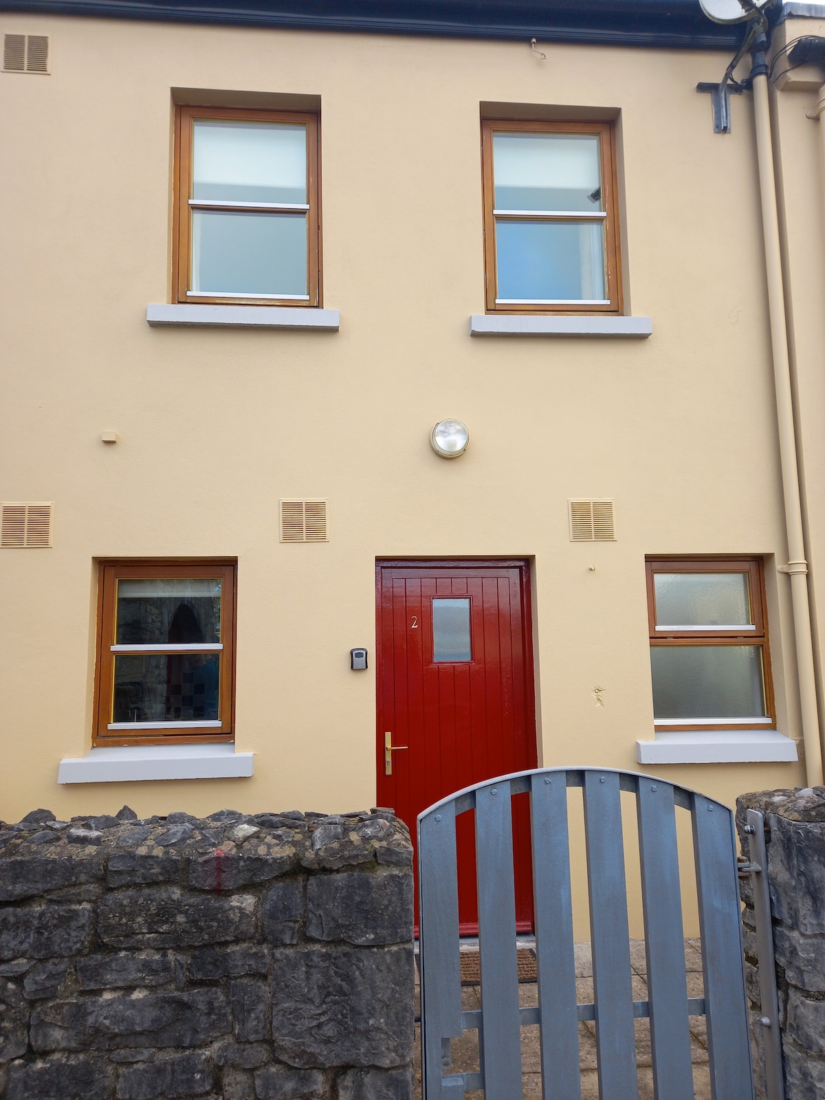 A two-story home is depicted, featuring a smooth beige exterior. A vibrant red front door is centered, flanked by two windows on the upper level and one window on the lower level. A stone wall leads to the gated entrance, offering a welcoming appearance.