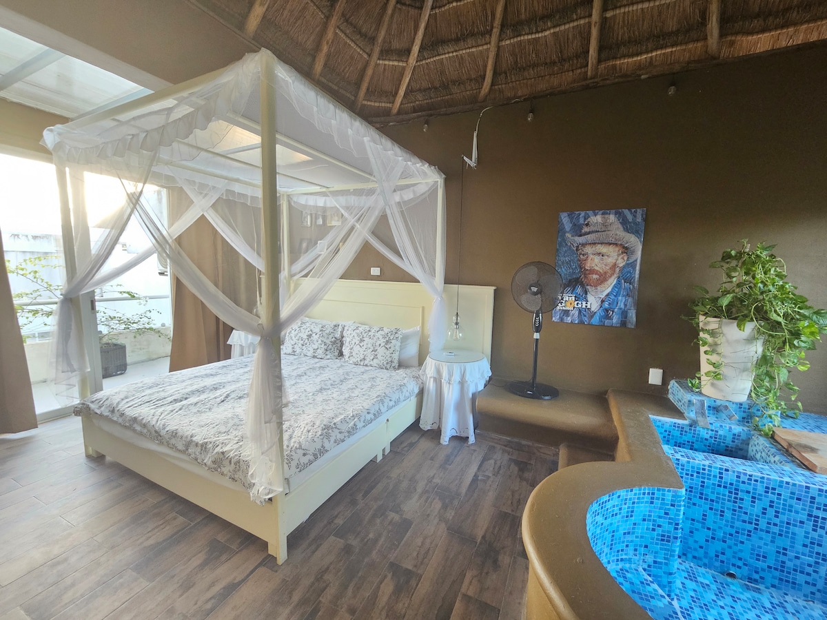 A spacious king suite is highlighted by a canopy bed draped in sheer white fabric. A lounge area features a cozy seating arrangement near a blue-tiled hot tub. Natural light filters in through large windows, complementing the earthy tones of the space.